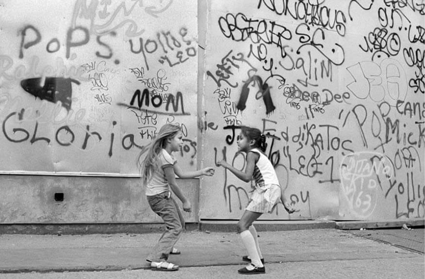 Historische Schwarz-Weiß-Aufnahme von zwei Kindern vor einer mit Tags besprühten New Yorker Wand, die die Anfänge des Graffiti in den 1970er-Jahren in New York symbolisiert. Auf der Wand ist der berühmte Tag „TAKI 183“ zu erkennen.