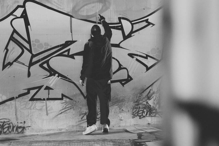 Schwarz-Weiß-Foto eines Graffiti-Künstlers von hinten, der mit einer Spraydose ein großes, komplexes Piece auf eine Wand sprüht.