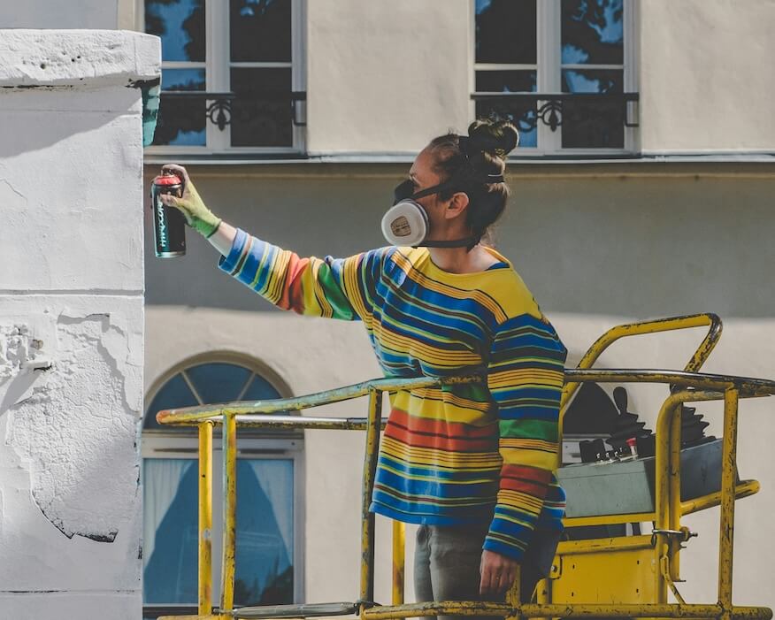 Eine Graffiti-Künstlerin auf einer Hebebühne, die eine Wand mit einer Sprühdose bemalt. Sie trägt eine bunte Jacke und eine Atemschutzmaske, was die professionelle Ausführung der Kunst betont.