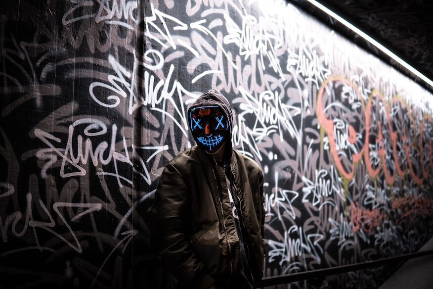 Eine Graffiti-Künstlerin auf einer Hebebühne, die eine Wand mit einer Sprühdose bemalt. Sie trägt eine bunte Jacke und eine Atemschutzmaske, was die professionelle Ausführung der Kunst betont.