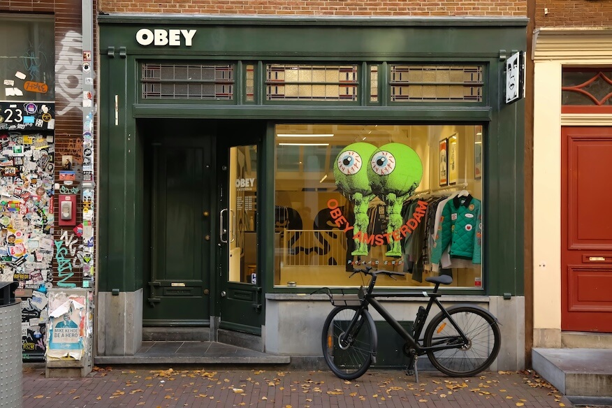 Außenaufnahme eines dunkelgrünen OBEY-Ladens in Amsterdam, mit großen Augen-Symbolen im Schaufenster und einem schwarzen Fahrrad davor.