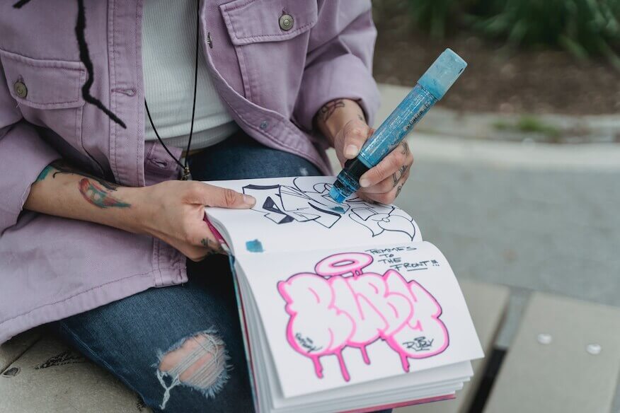 ine Person zeichnet mit einem blauen Marker einen pinken Graffiti-Entwurf in ein Skizzenbuch. Ihre Hände sind mit Tattoos bedeckt.