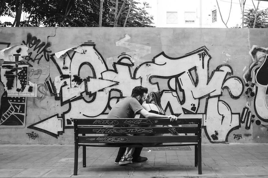 Schwarz-Weiß-Foto eines Paares, das auf einer Parkbank vor einer Wand mit klassischen Graffiti-Schriftzügen sitzt.