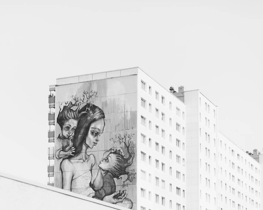 Ein großflächiges, schwarz-weißes Mural von Herakut auf der Seite eines modernen Wohngebäudes. Das Kunstwerk zeigt abstrakte Figuren und Bäume und steht im Kontrast zur klaren Architektur des Gebäudes.