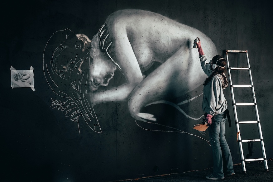 Künstlerin Hera sprüht auf eine dunkle Wand und arbeitet an einem großen, grauen Mural, das eine liegende Figur darstellt. Eine Leiter und eine Skizze der Figur sind zu sehen.
